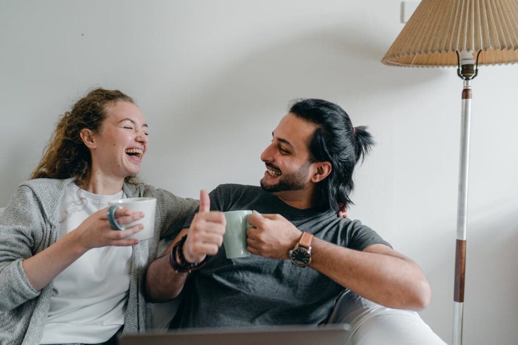 A man and woman sit together on a couch, smiling and holding mugs. The woman is laughing while the man gives a thumbs up. A lamp is positioned next to them.