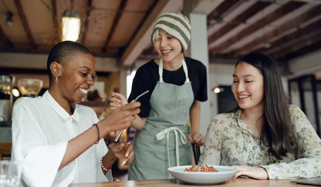 Three people, appearing to be of lower middle class, are sitting at a table; one is holding utensils and smiling, another is wearing an apron and beanie standing, and the third is seated with a bowl of food in front of her, also smiling.