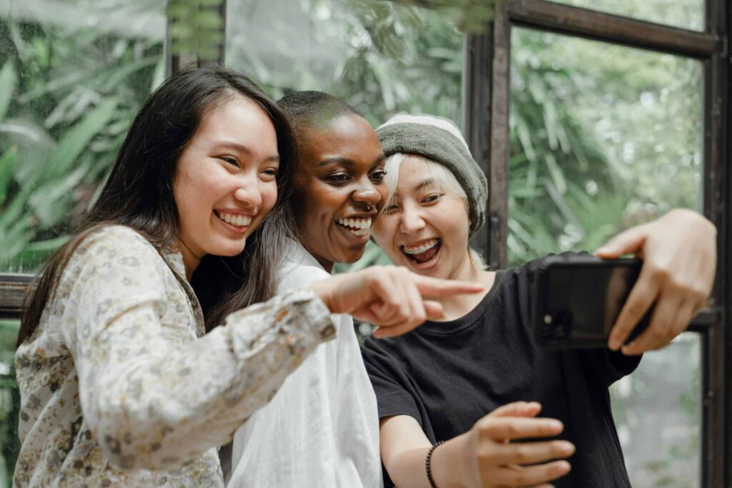 Three people are standing close together, smiling, and taking a selfie indoors with greenery visible through the windows behind them, capturing a moment that will soon be shared on social media.