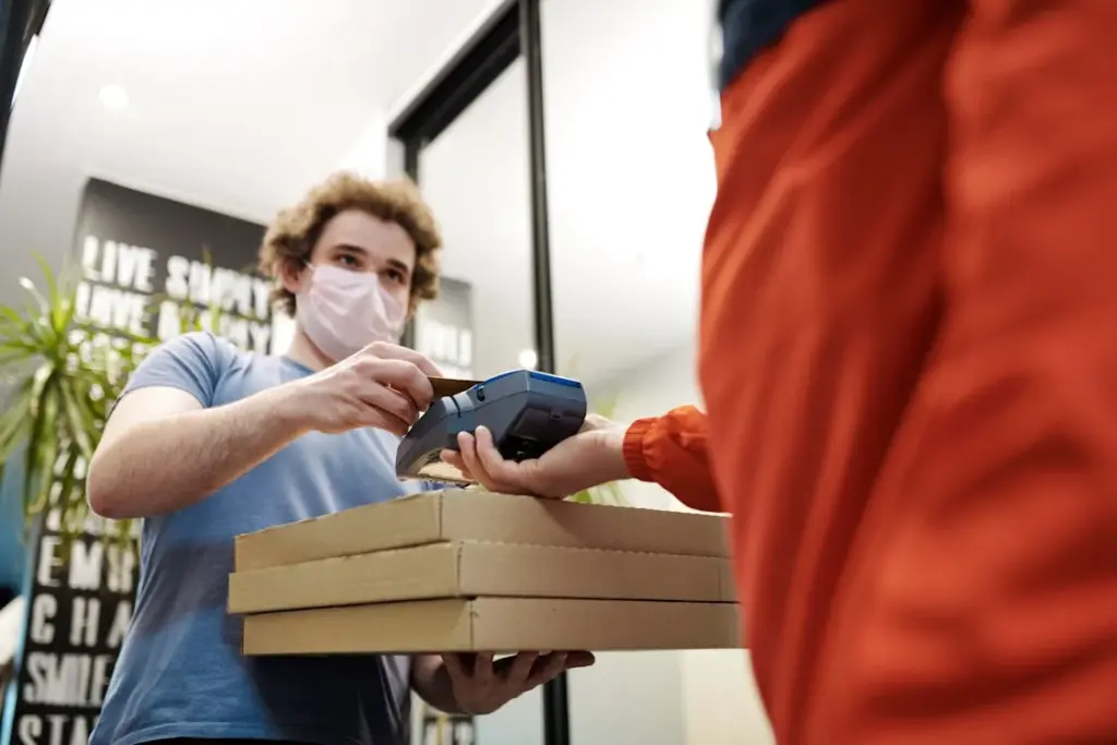 A person in a face mask holds pizza boxes while processing a payment from another person dressed in red, a reminder that some consider pizzerias as places where it is not recommended to leave a tip.
