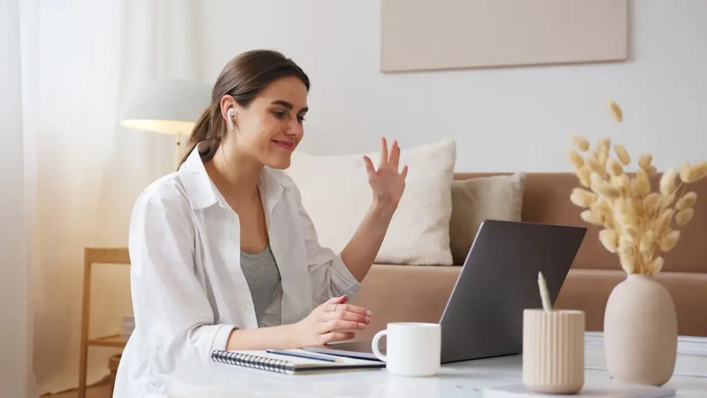 A woman with earbuds waves at a laptop screen while sitting at a table with a notebook, pen, and coffee mug in a cozy living room, surrounded by things replaced by technology.