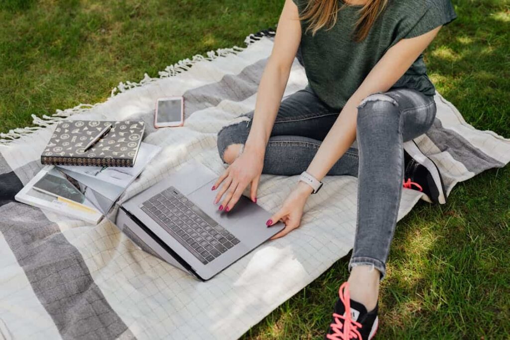 Person sitting on a striped blanket outdoors, using a laptop. Nearby are a smartphone, a notebook with a pattern, and documents—reminders of the many things replaced by technology.