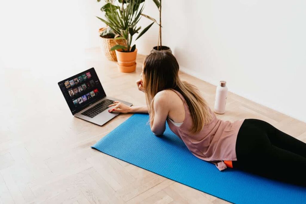 A woman in sportswear lies on a blue yoga mat, watching media on a laptop. A water bottle is next to her, with plants in the background—signs of a world where even relaxation routines are influenced by things replaced by technology.