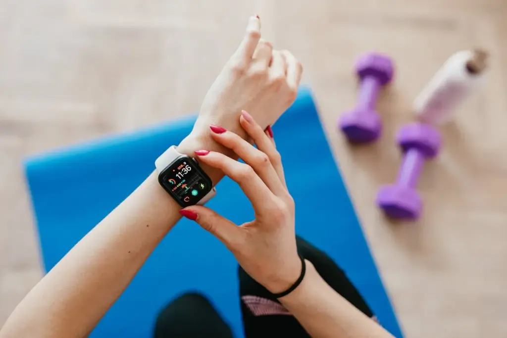 A person adjusts a smartwatch on their wrist while sitting on a blue exercise mat. Purple dumbbells and a water bottle are visible in the background.