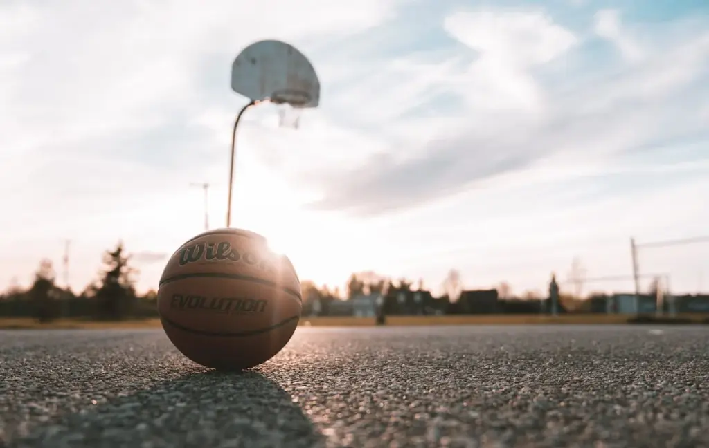 A basketball lies on an outdoor court with a hoop in the background under a clear sky, capturing a simple moment that resonates deeply with the working class.