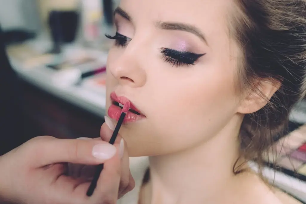Makeup artist applies pink lip gloss to a woman's lips as she closes her eyes, highlighting her winged eyeliner and shimmering eyeshadow.