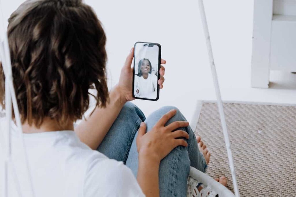 A person with short hair is sitting on a chair, holding a smartphone and engaged in a video call, highlighting how many things have been replaced by technology.