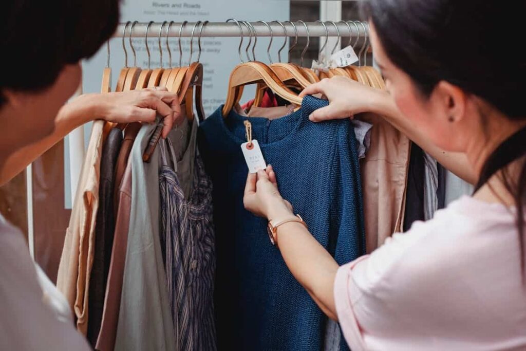 Two people with a lower middle class income examine a blue dress on a wooden hanger, checking the price tag, in a clothing store. More clothing items are hanging on the rack.