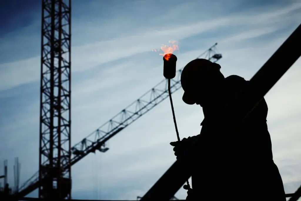 A silhouette of a construction worker holding a lit blowtorch against the background of cranes and an industrial skyline embodies the essence of the working class.