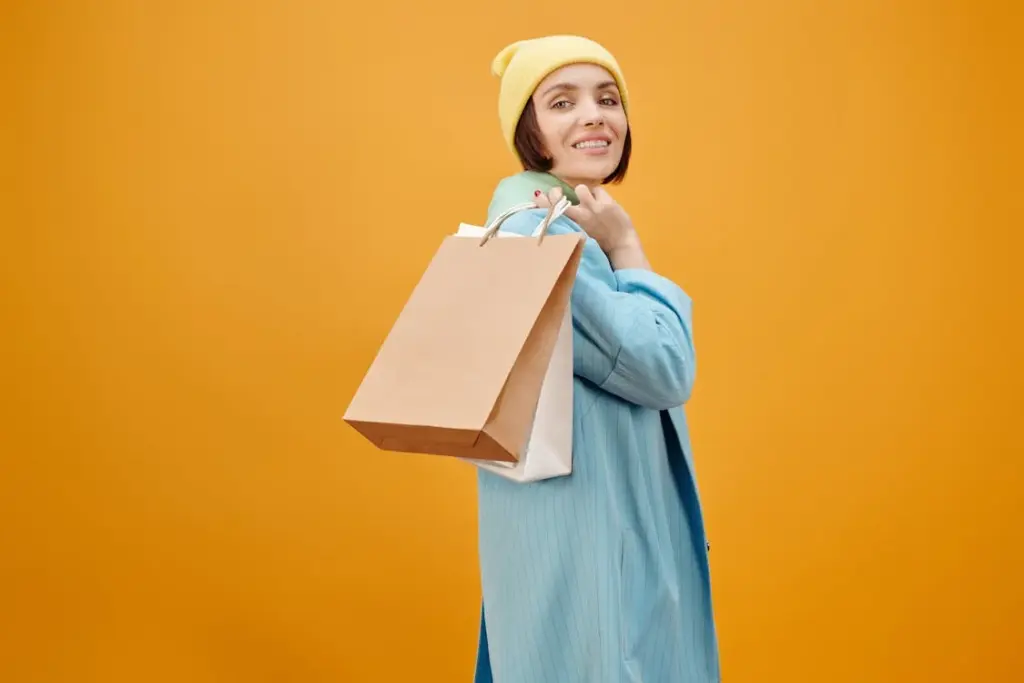 Person in a light blue coat and yellow beanie holding shopping bags over their shoulder, standing in front of an orange background.