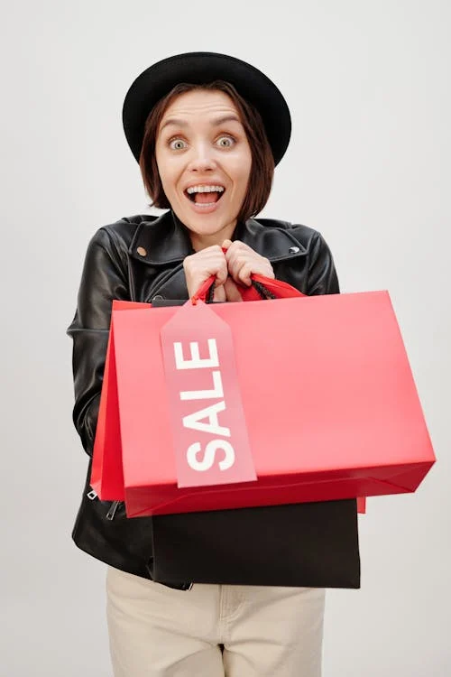 Person with an excited expression, wearing a hat and black jacket, holding large red shopping bags with a "SALE" tag.