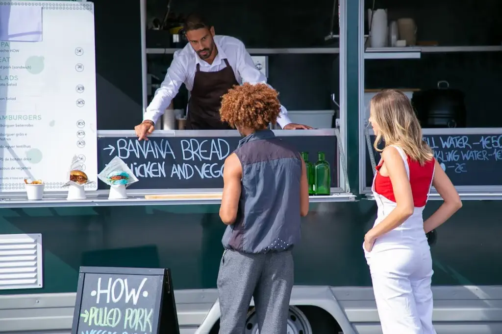 Two customers stand in front of a food truck, interacting with a server. A menu and food items are visible on the counter. The male server is pointing to a chalkboard menu, at this place not to tip.