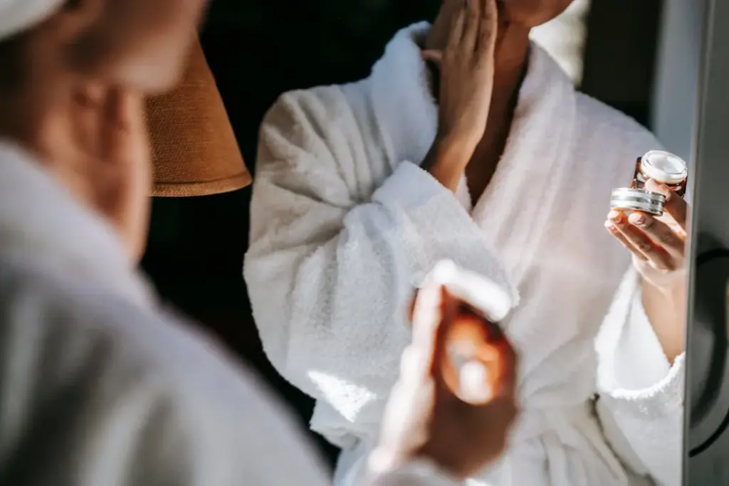 Person in a white bathrobe applying face cream while looking in a mirror, holding a small jar of the cream with one hand.