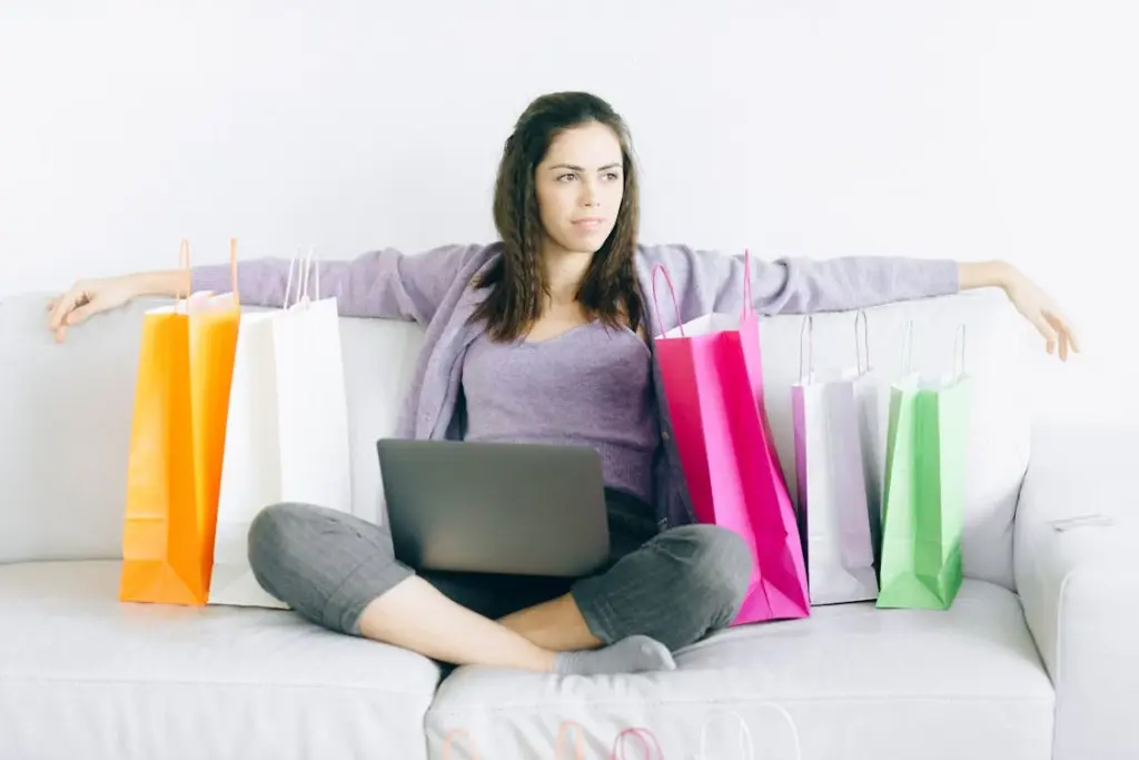 A woman sits on a couch with several shopping bags around her. She has a laptop on her lap and looks to the side with a thoughtful expression.
