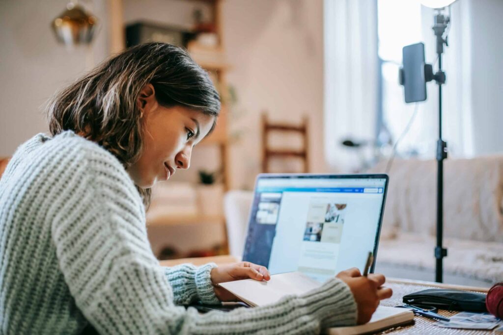 A person writes in a notebook while looking at a laptop screen in a home setting, pondering the many things replaced by technology.