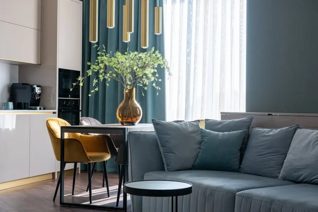 Modern living room with a gray sectional sofa, a round black coffee table, a yellow chair at a small dining table, a vase with green branches, and pendant lights. Kitchen appliances in the background.