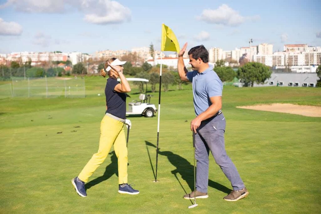 Two golfers, evidently enjoying their upper class income, exchange a high-five near a flag on a pristine golf course under a clear sky, with an elegant golf cart and the cityscape in the background.