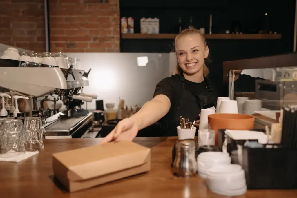 A smiling barista stands behind a coffee shop counter, handing a takeout box to a customer. Coffee equipment and utensils are visible in the background, embodying the hard-working spirit of the working class.