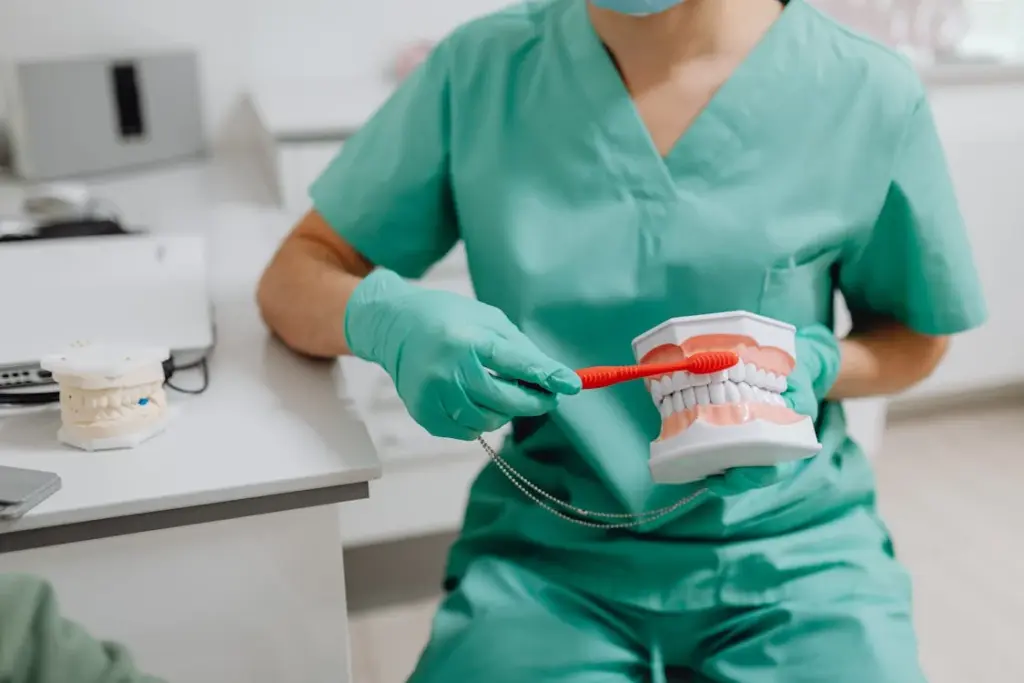 A dental professional in green scrubs demonstrates brushing technique on a dental model with a red toothbrush.