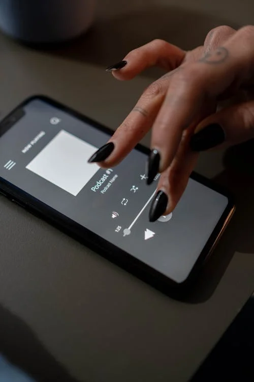 A person with black nail polish adjusts the volume on a podcast playing on a smartphone, reflecting how many things replaced by technology have seamlessly integrated into our daily routines.