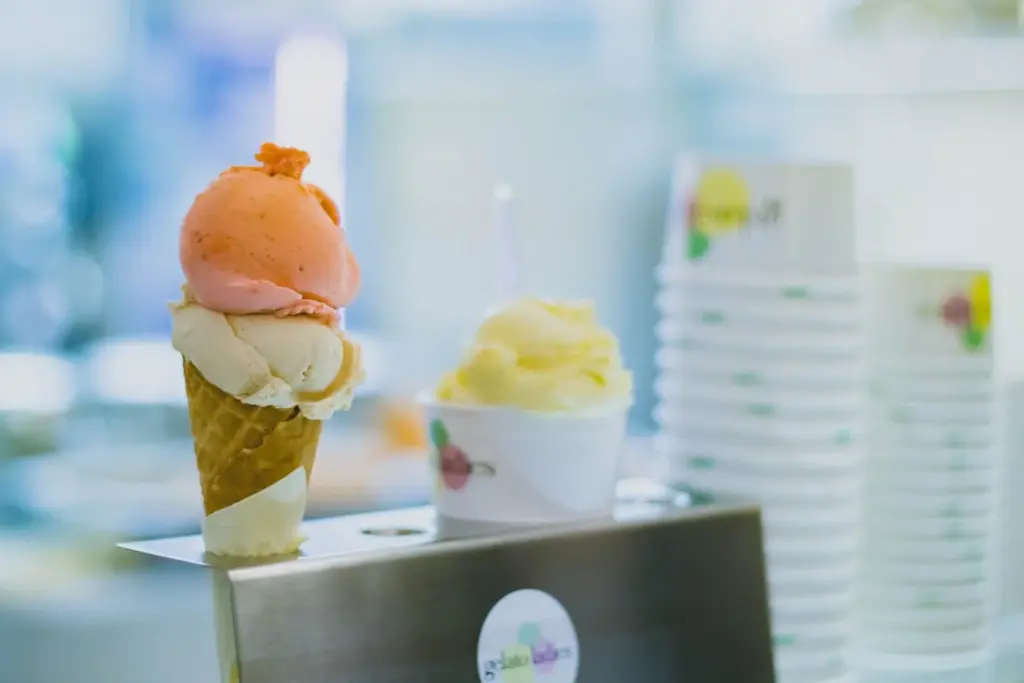 A strawberry and vanilla ice cream cone and a cup of vanilla ice cream are placed on a counter next to a stack of empty cups in a place not to tip.
