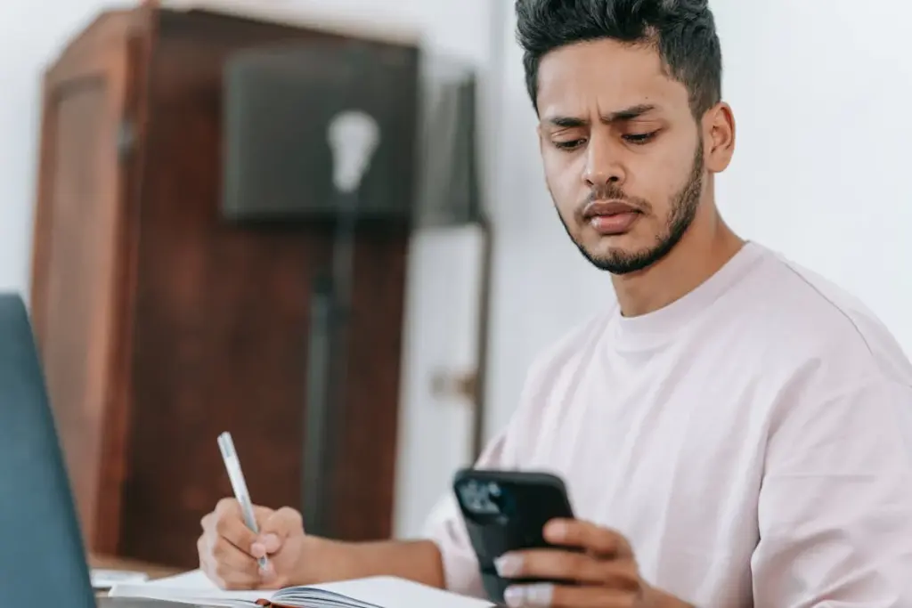 A person holding a phone in one hand while writing with a pen in the other, looking at the phone screen with a concerned expression. A laptop is open in front of them on the table, highlighting the tension between traditional tools and things replaced by technology.