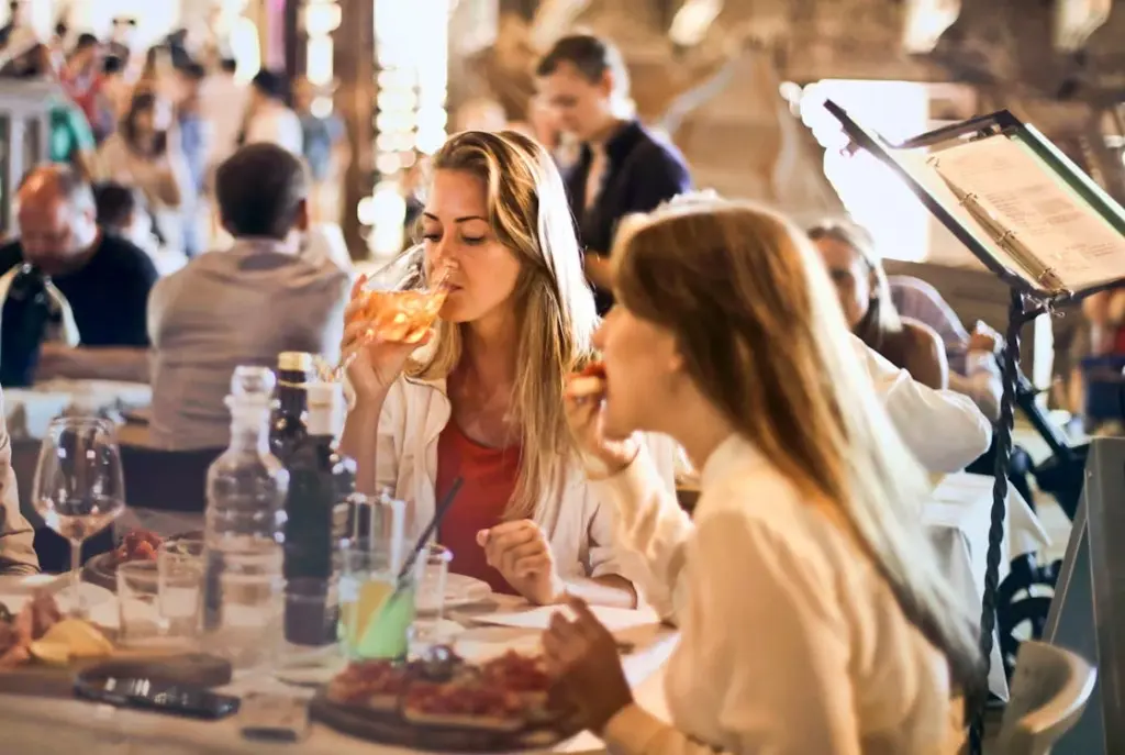 Two women are seated at a restaurant table, enjoying their meal and drinks, exuding an air of upper-class sophistication. In the background, other diners and a server are visible, painting a picture of refined dining.