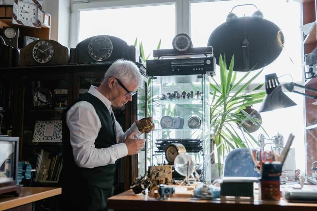 An older man with white hair inspects a small clock in a cluttered workshop filled with various clocks and tools, one of those places where it is not recommended to leave a tip.