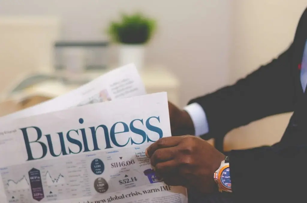 A person from the upper class reads a newspaper with the headline "Business" and a table of financial data in the background. A wristwatch and suit sleeve are visible on their arm holding the paper, reflecting their upper class earnings.