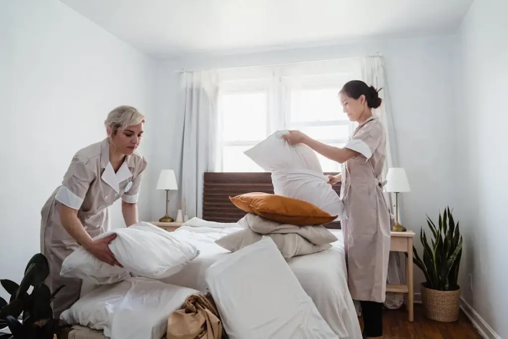 Two women, perhaps weary from the pervasive tip fatigue many Americans experience, are making a bed in a brightly lit room with white walls and large windows. One woman is arranging pillows while the other is adjusting the sheets.