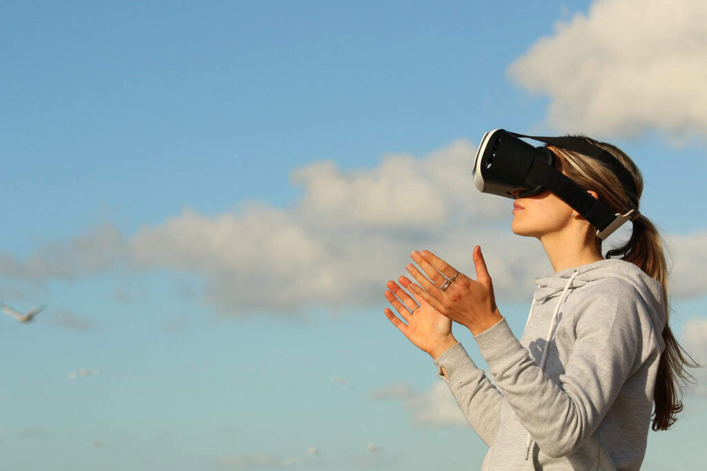 A person wearing a gray hoodie and VR headset gestures with their hands against a blue sky with scattered clouds and flying seagulls, symbolizing how things replaced by technology continue to shape our interaction with the world.
