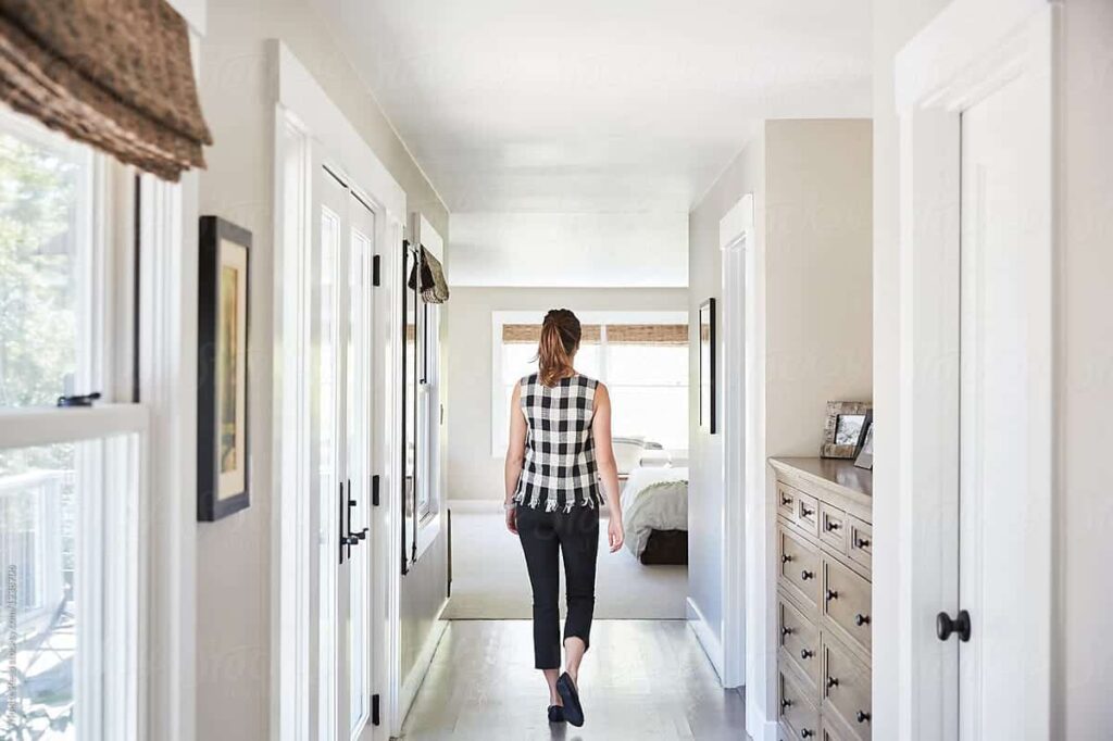 A woman in a checkered top walks down a bright hallway towards a bedroom. The room has light-colored walls, white doors, and a chest of drawers on the right.