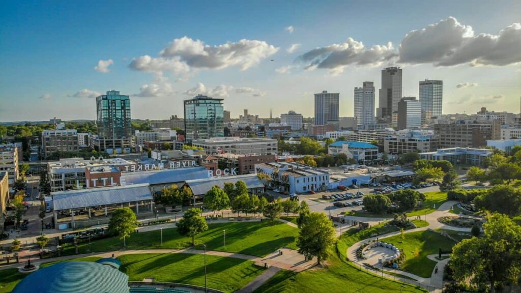 Aerial view of Little Rock, Arkansas, one of the most comfortable cities to live in the US, showcasing a mix of modern high-rises and historic buildings, with lush green parks and a clear sky with scattered clouds.