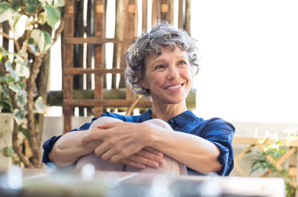 A woman with short curly gray hair sits outdoors, smiling, with her arms resting on her knees. A wooden trellis and greenery are visible in the background.