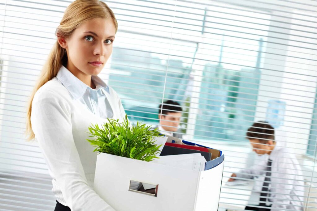A woman with a serious expression holds a box containing office supplies and a plant. Two people in office attire are visible in the background through blinds.