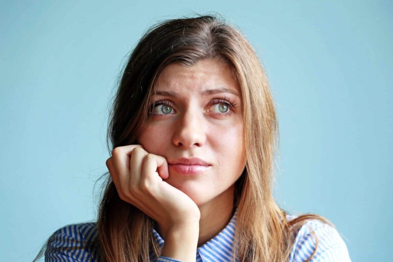 A woman with long brown hair rests her chin on her hand, looking upwards with a thoughtful expression against a light blue background. She is wearing a striped shirt.