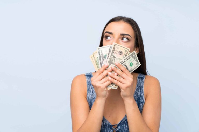 A woman in a blue dress holds a fanned-out stack of U.S. dollar bills in front of her face, looking to her right against a light blue background.