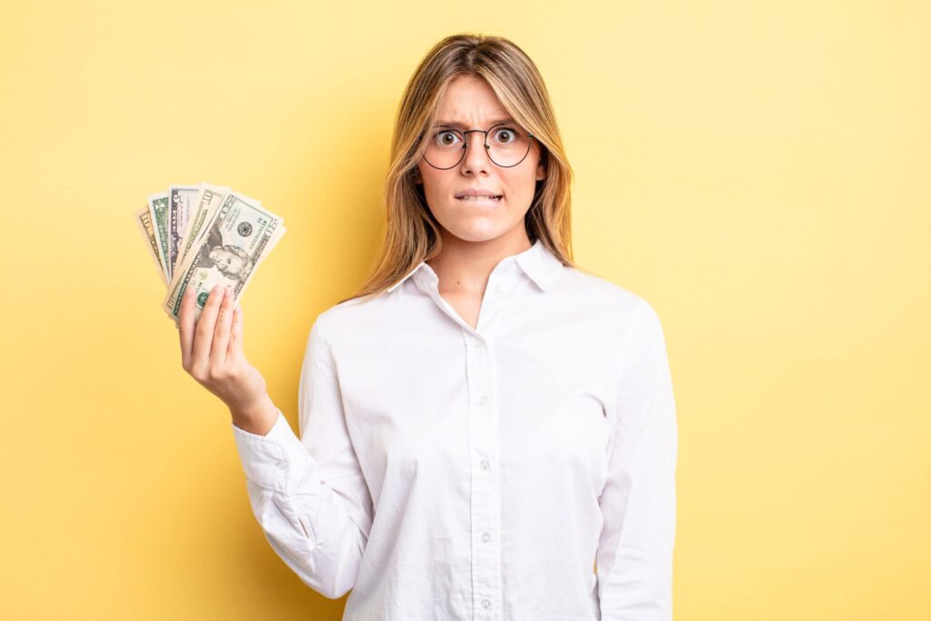A person with long hair and glasses holds a fanned-out stack of U.S. dollar bills, looking concerned against a yellow background.