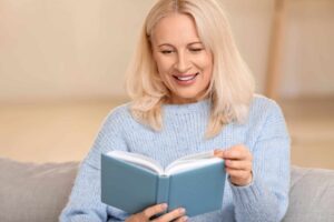 A woman with blonde hair and a light blue sweater is sitting on a couch, smiling, while reading a book with a blue cover.