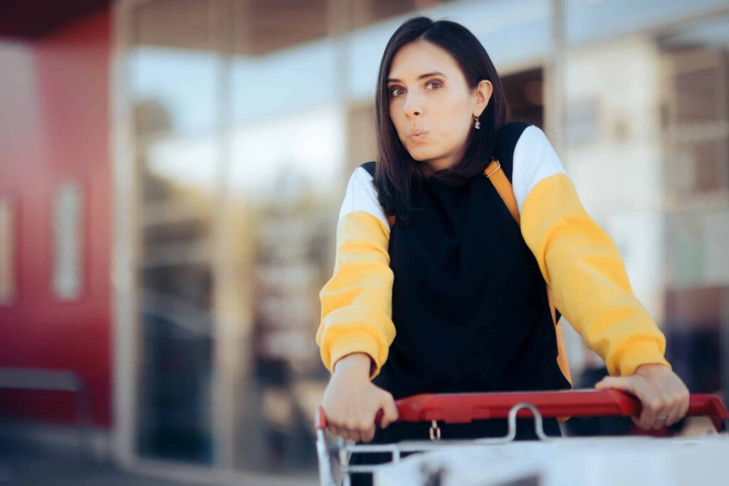 A woman wearing a yellow and black hoodie stands outdoors, leaning on a shopping cart, with an expression of mild surprise or contemplation.