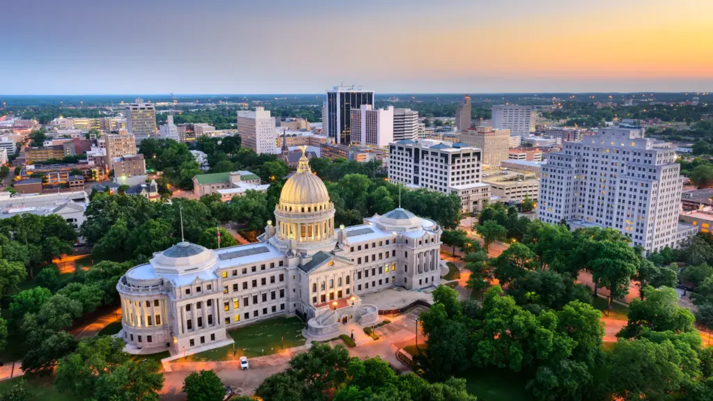 Aerial view of a cityscape at dusk with a prominent government building featuring a dome in the foreground, surrounded by numerous other buildings and greenery. This picturesque setting exemplifies one of the most comfortable cities to live in the US, bathed in the warm hues of the setting sun.