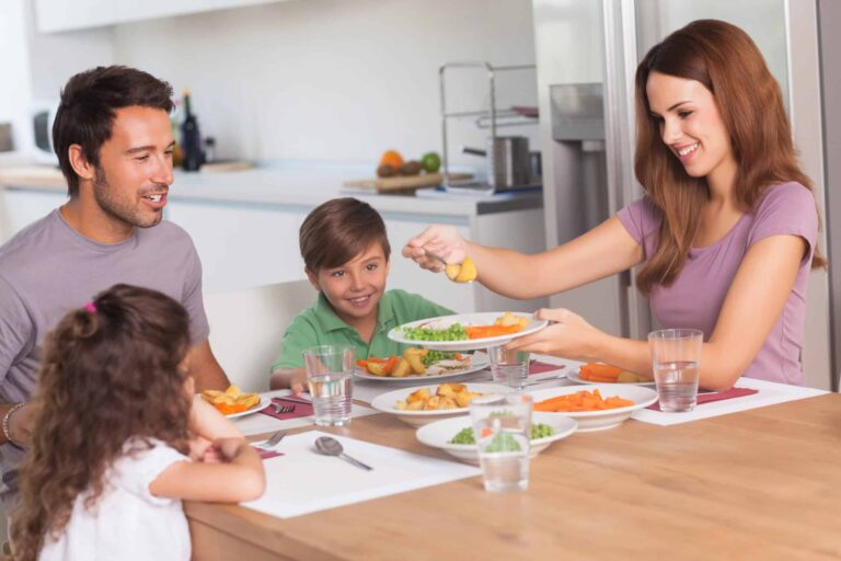 A family of four sits at a dining table, smiling and enjoying a meal together. The table is set with plates of food, glasses of water, and cutlery. The mother serves food from a large dish.