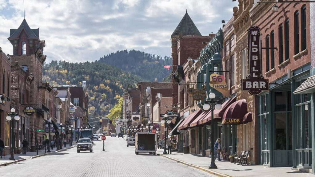 A street with historic buildings and signage, including "Wild Bill Bar," lined with trees and hills in the background, under a partly cloudy sky. It's scenes like these that make some of America’s most comfortable cities to live in US so charming and desirable.