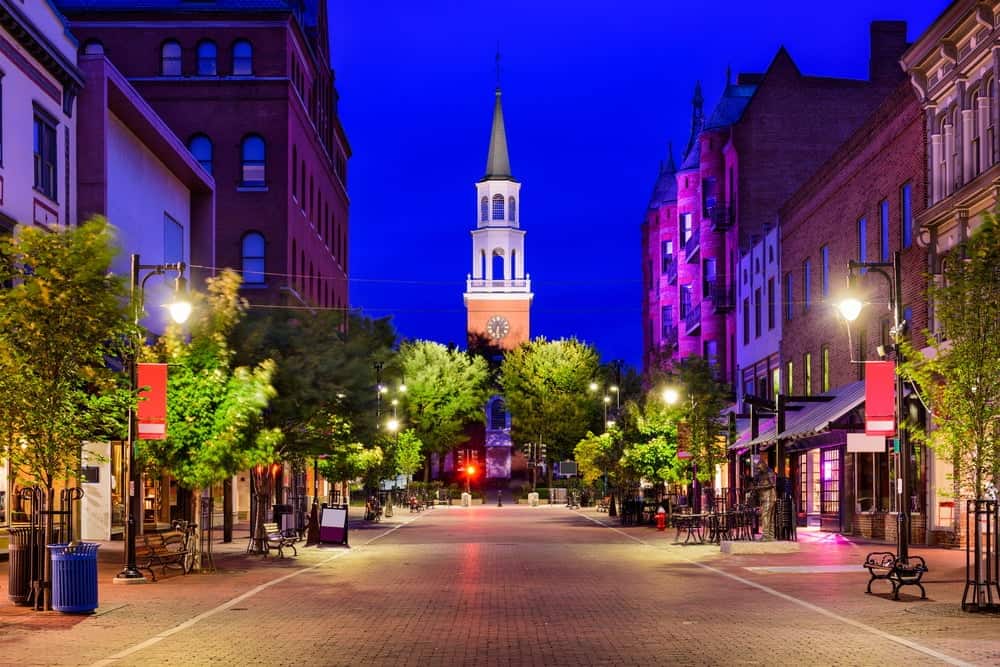 A well-lit pedestrian street lined with trees and buildings leads to a clock tower with a spire, under a deep blue evening sky in one of the most comfortable cities to live in the US.