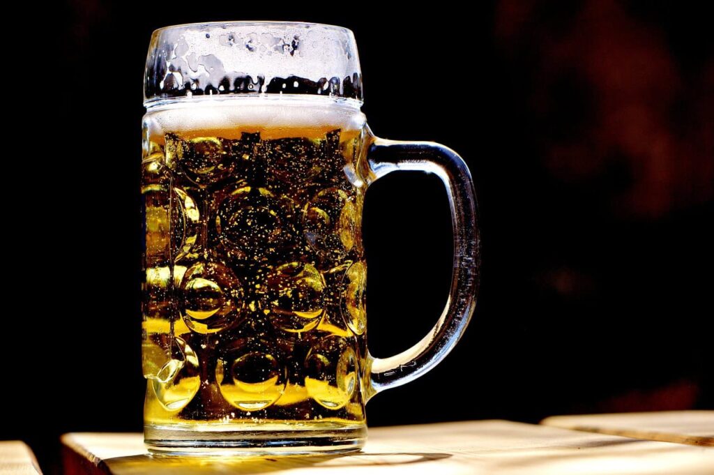 A clear glass mug filled with frothy beer sits on a wooden surface against a dark background.