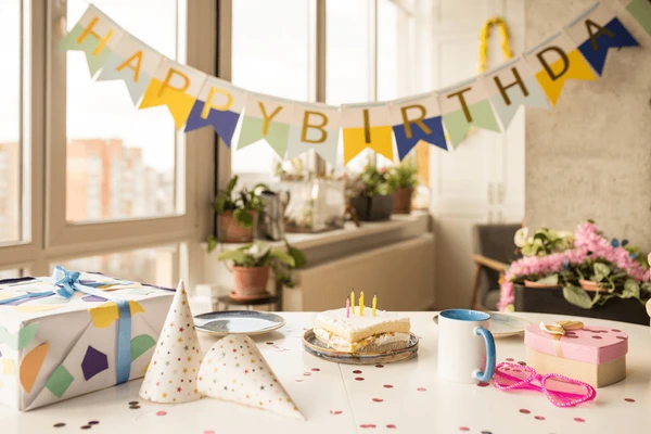 A table with birthday decorations including a "HAPPY BIRTHDAY" banner, a wrapped gift, cone hats, a cake with candles, and various other decorative items.