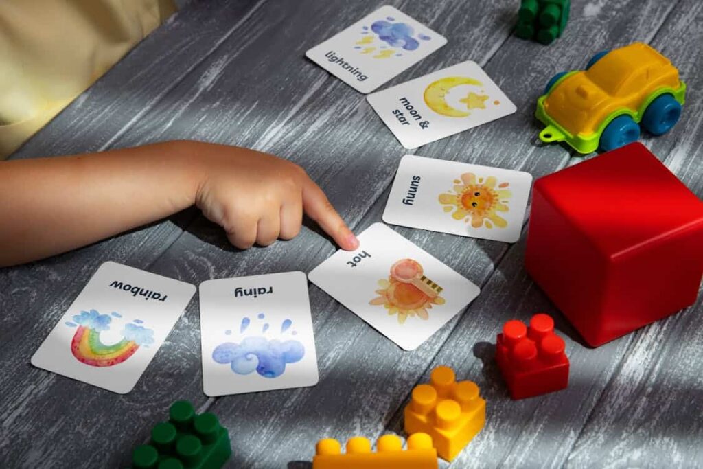 A child's hand points to a card with "hot" written on it, surrounded by other cards, building blocks, and a toy car on a textured gray surface.