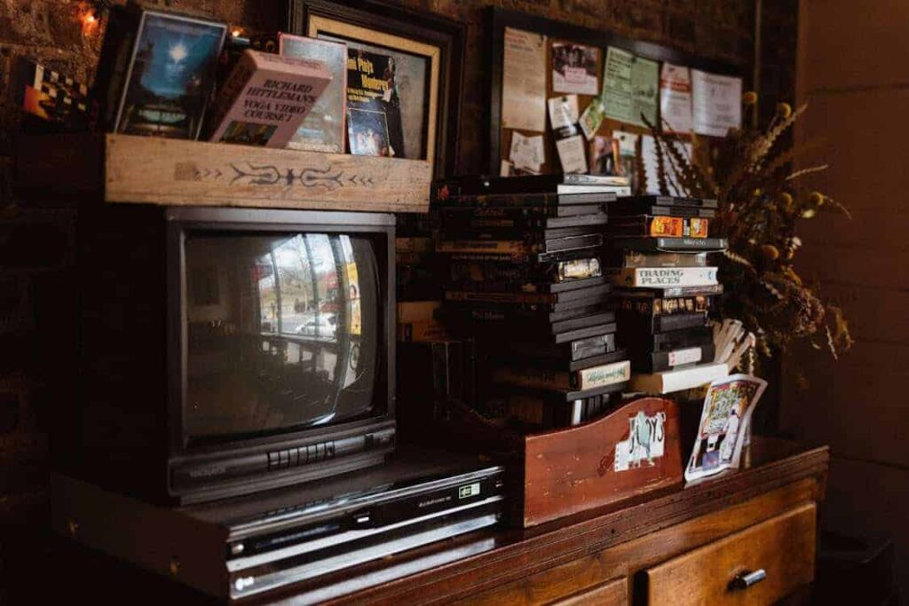 A vintage TV and VHS player sit on a wooden cabinet, surrounded by stacks of VHS tapes, framed pictures, and a bulletin board on a brick wall.