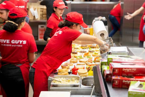 Workers in red Five Guys uniforms prepare burgers and fries in a busy kitchen. Various toppings and buns are arranged on the counter.