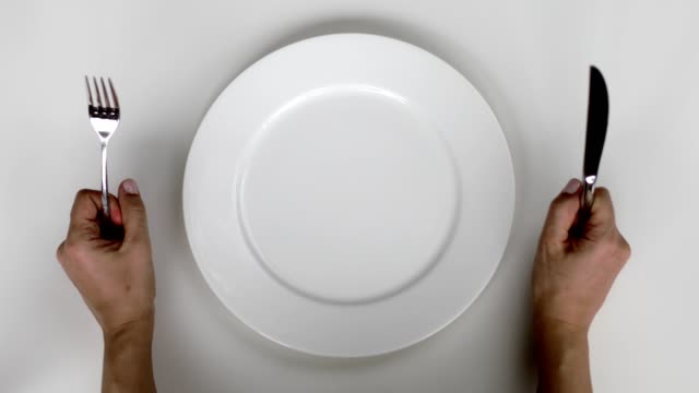 A person's hands holding a fork in the left hand and a knife in the right hand, positioned on either side of an empty white plate on a white background.